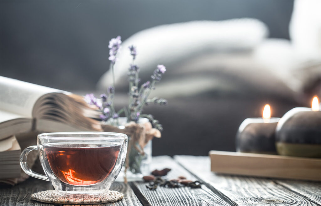 Rauhoittava asetelma, jossa teekuppi, avonainen kirja, kynttilöitä ja laventelia pölydällä
